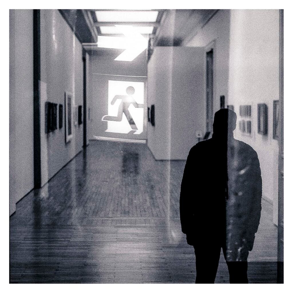 Silhouette superposée à un couloir du Musée d’Art avec un pictogramme de sortie, en double exposition réalisée au Nikon F3 sur film Fomapan 400.