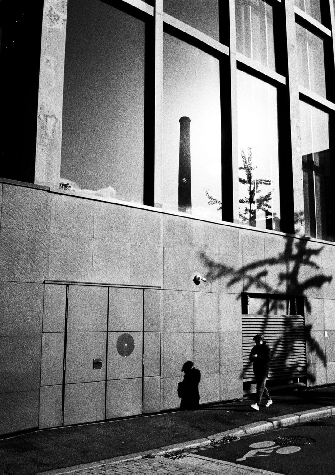 Reflet d’une cheminée industrielle dans la façade vitrée d’un bâtiment de l’Île de Nantes, avec ombre d’un arbre et silhouette d’un passant.