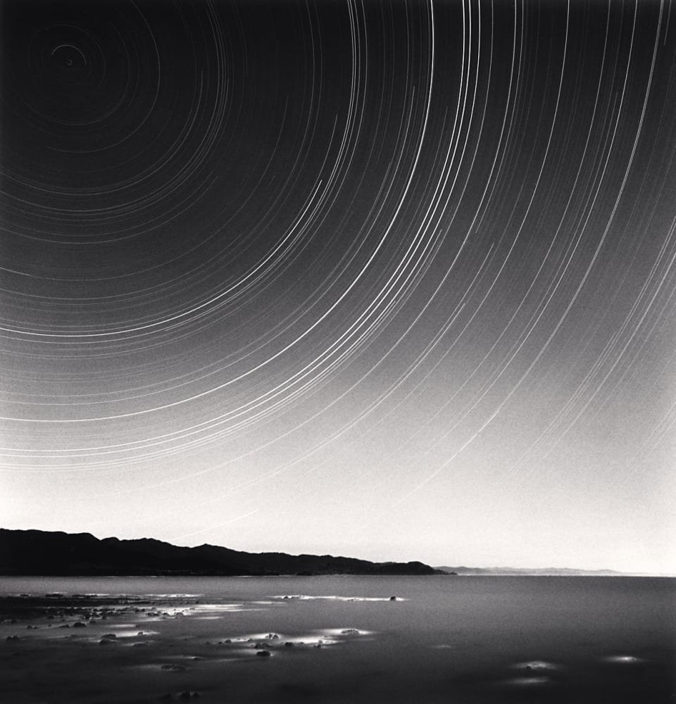 Traînées d’étoiles formant de larges arcs circulaires au-dessus du littoral de Te Kaha, photographiées en longue pose par Michael Kenna en 2014.