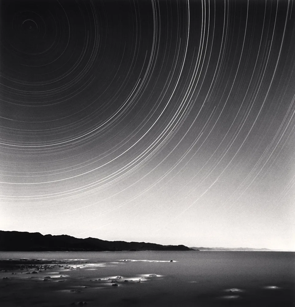Traînées d’étoiles formant de larges arcs circulaires au-dessus du littoral de Te Kaha, photographiées en longue pose par Michael Kenna en 2014.
