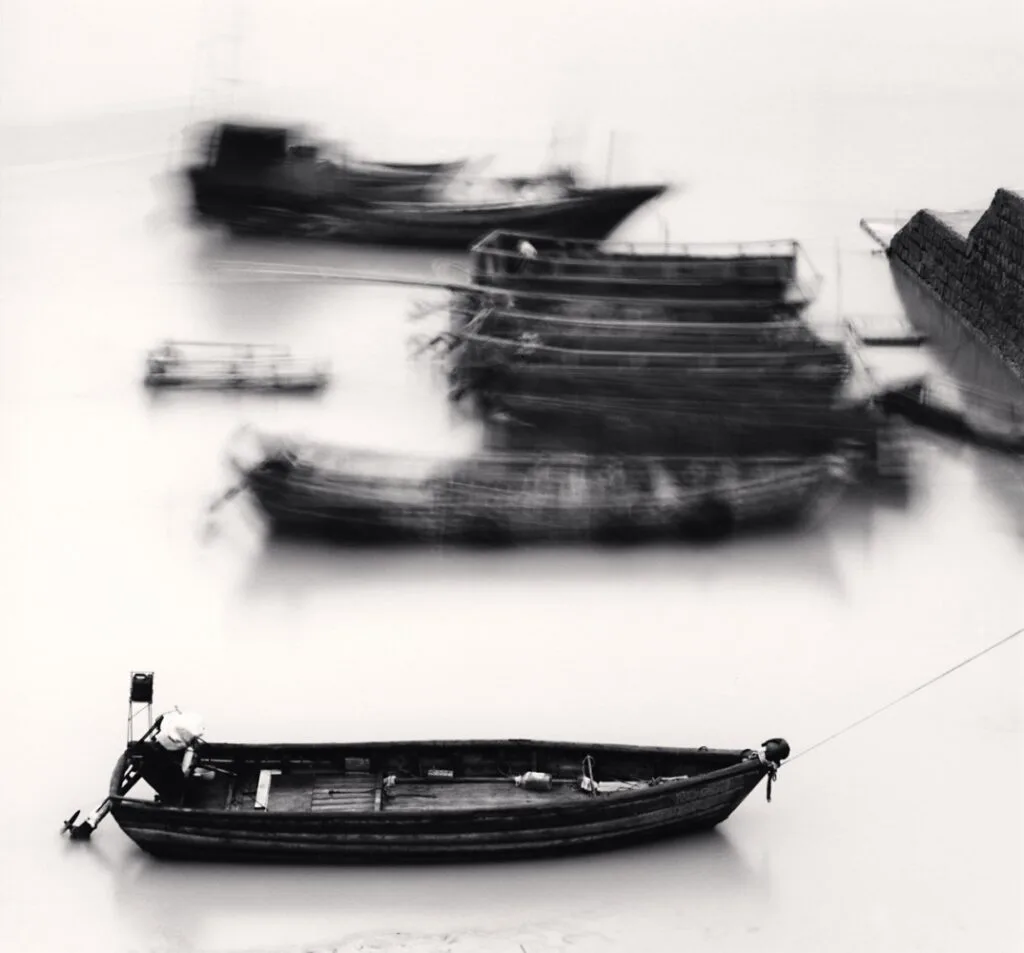Barque amarrée au premier plan avec bateaux flous en arrière-plan, photographiée en noir et blanc à Xiapu par Michael Kenna en 2010.