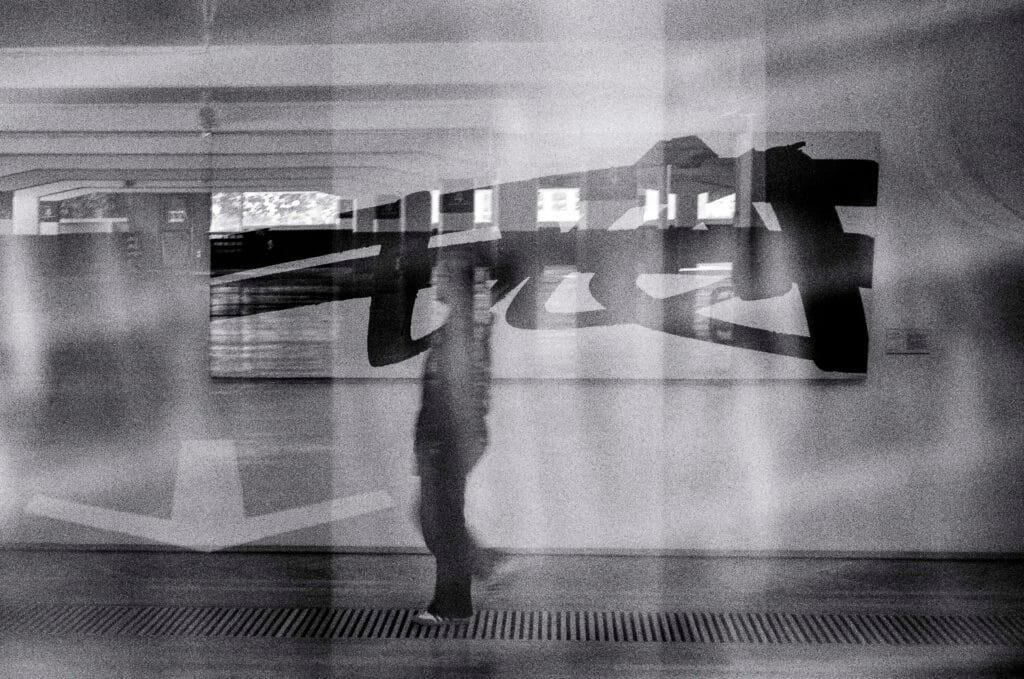 Superposition accidentelle entre un tableau contemporain du Musée d’Art de Nantes et une silhouette floue dans le parking Gare Nord, en noir et blanc.