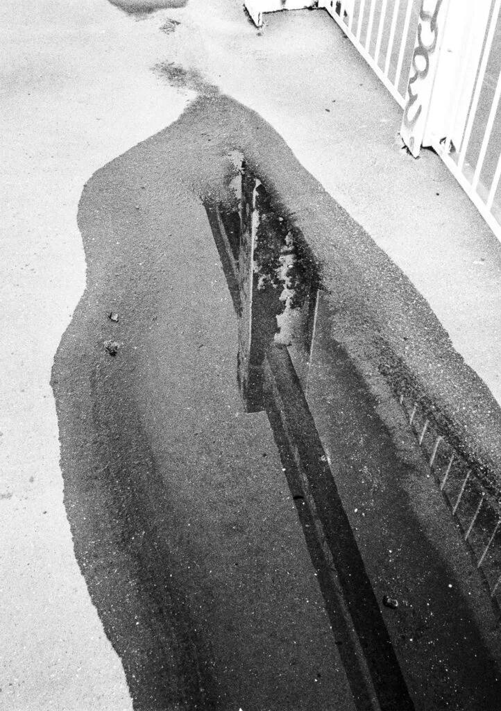 Reflet d’un bâtiment dans une flaque sur le bitume à Nantes, photographié en noir et blanc avec une pellicule périmée.