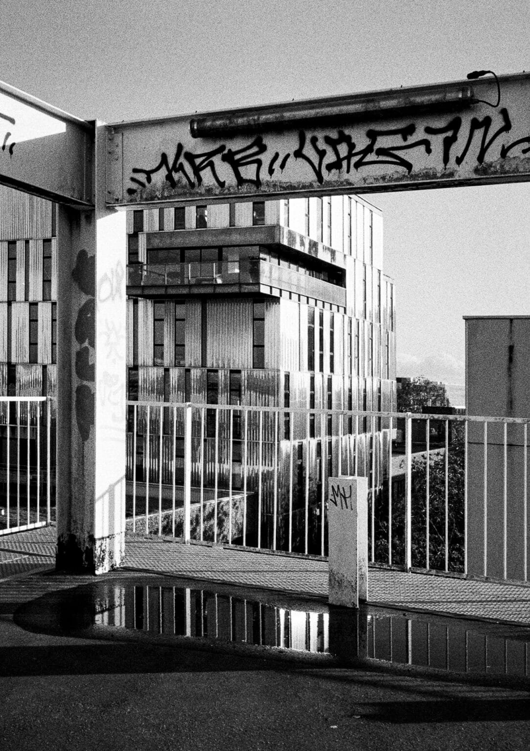 Immeuble moderne en façade métallique reflété dans une flaque d’eau sur le toit d’un parking, avec barrière taguée au premier plan.