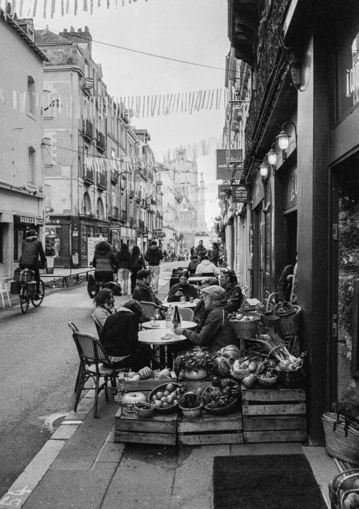 Terrasses animées un samedi midi rue Maréchal Joffre à Nantes, avec étals de légumes, passants, cyclistes et guirlandes suspendues.