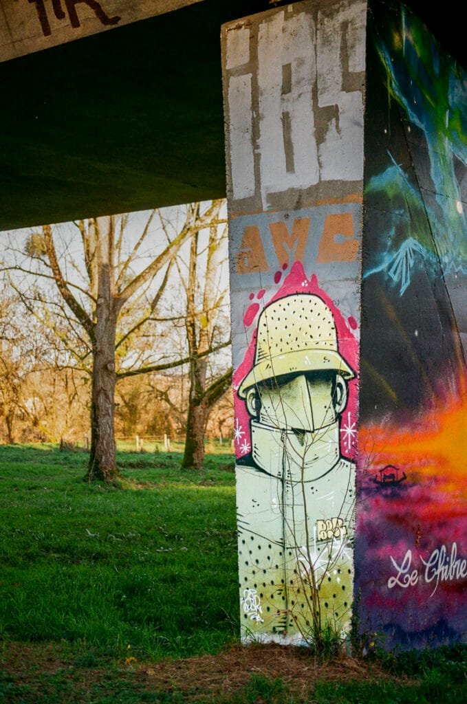 Graffiti représentant une silhouette casquée sur un pilier de béton sous un pont à Pirmil, dans un paysage arboré.