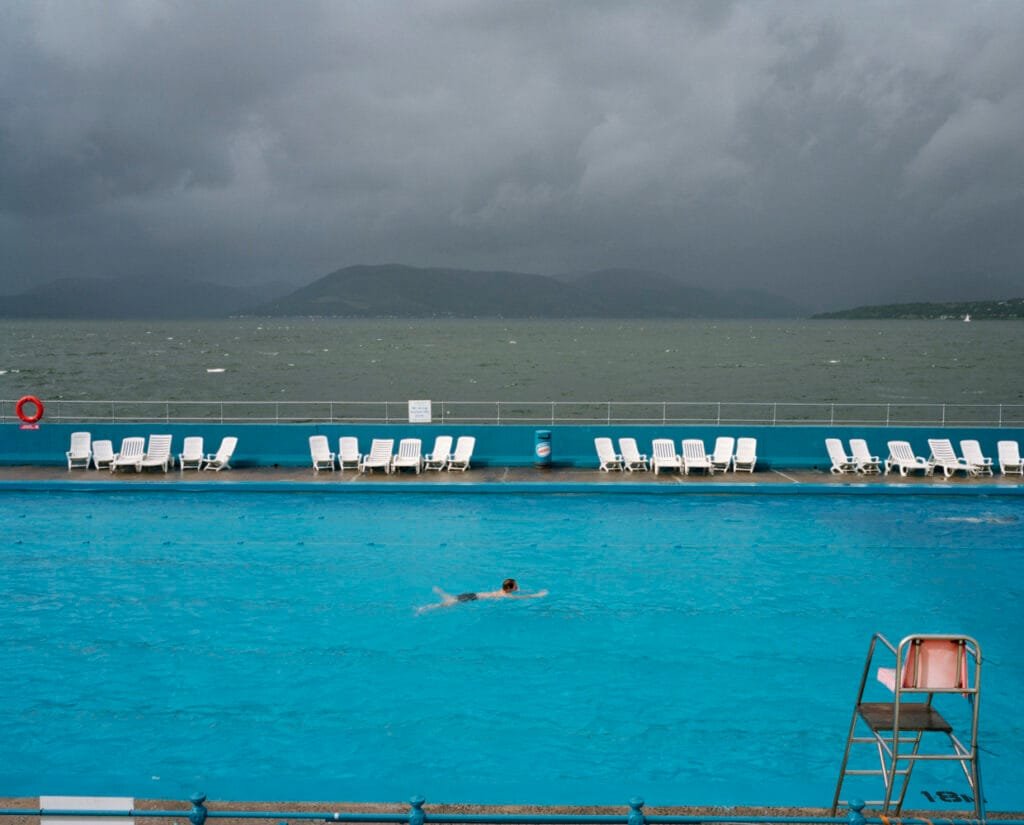 Piscine extérieure du Gourock Lido en Écosse sous un ciel orageux, avec un nageur solitaire. Photographie de Martin Parr en 2004.