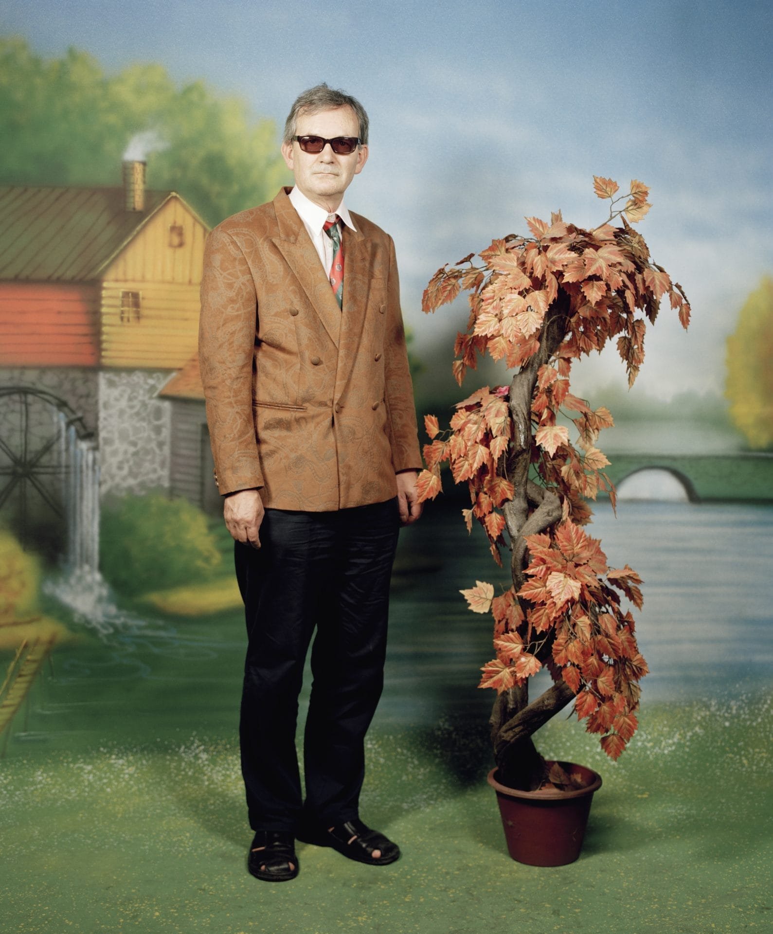 Portrait de Martin Parr posé dans un studio kitsch avec une plante artificielle, photographié dans un décor peint.