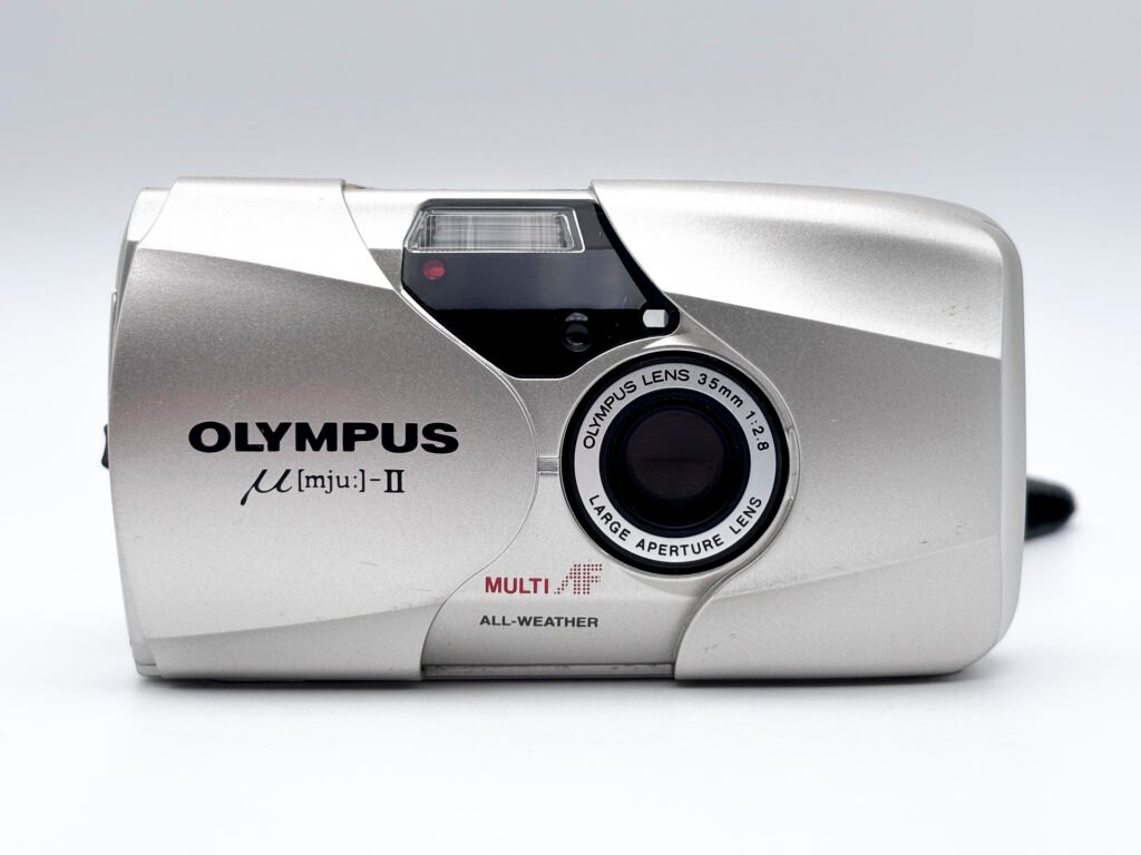 Olympus Mju II vu de face avec coque coulissante ouverte et objectif 35mm f/2.8 visible.