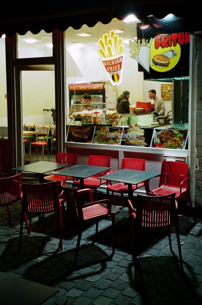 Snack éclairé de nuit place Sainte-Catherine à Bruxelles avec tables et chaises rouges en extérieur.