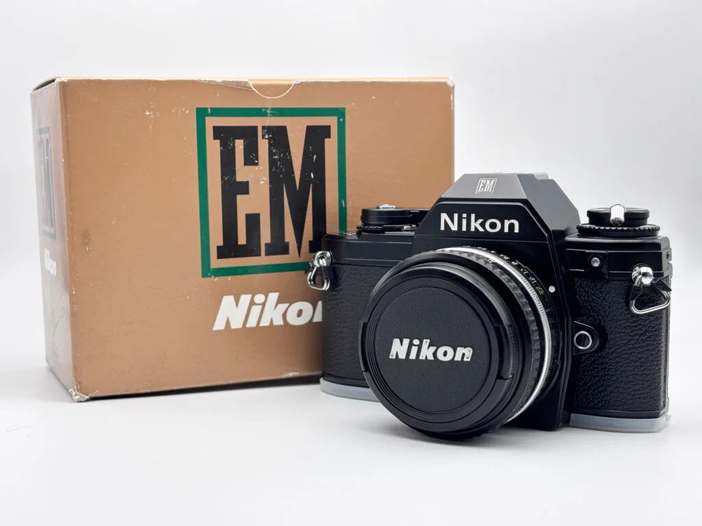 Nikon EM posé devant sa boîte d’origine sur fond neutre, reflex argentique compact de 1979.
