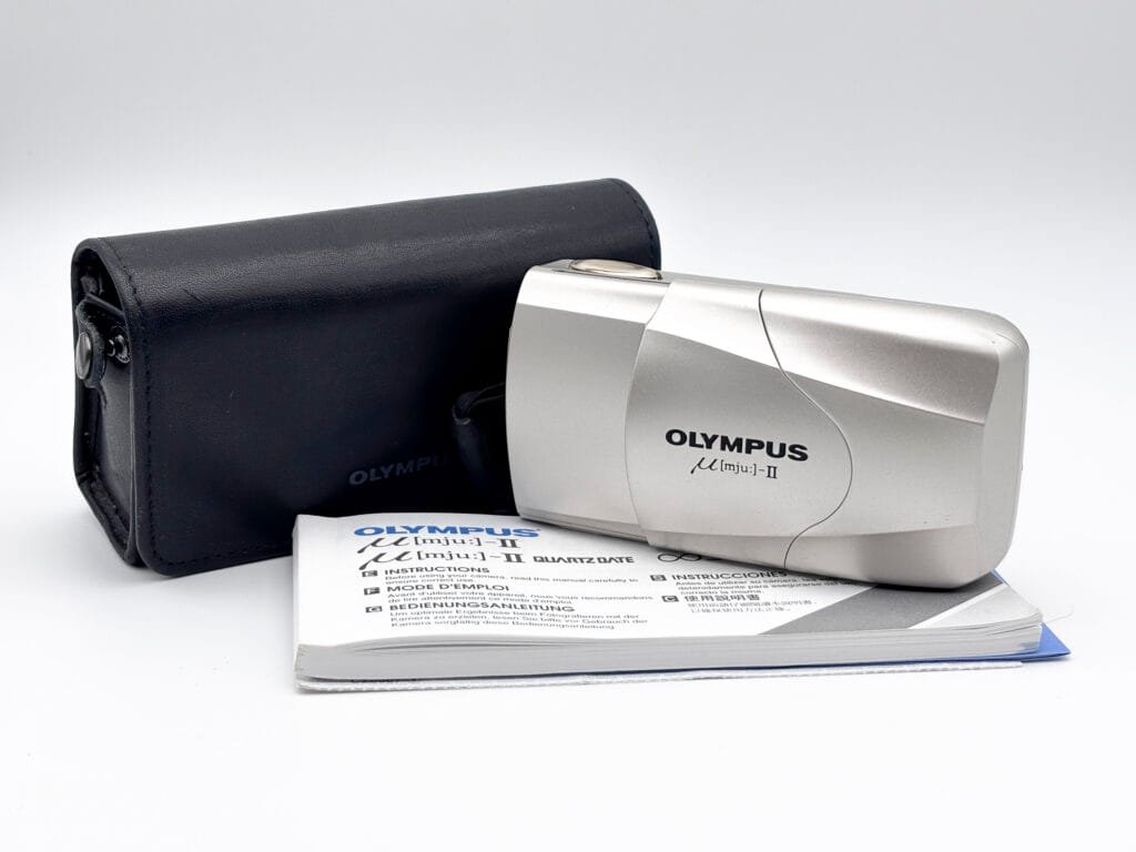 Olympus Mju II argent posé devant sa housse noire d’origine et son manuel d’instructions.