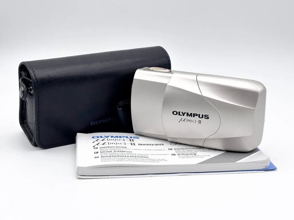 Olympus Mju II argent posé devant sa housse noire d’origine et son manuel d’instructions.