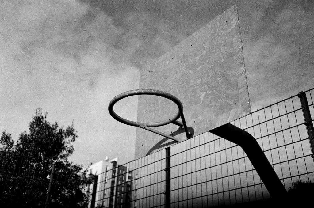 Panier de basket extérieur sans filet, photographié en contre-plongée, avec un grillage et des immeubles en arrière-plan.