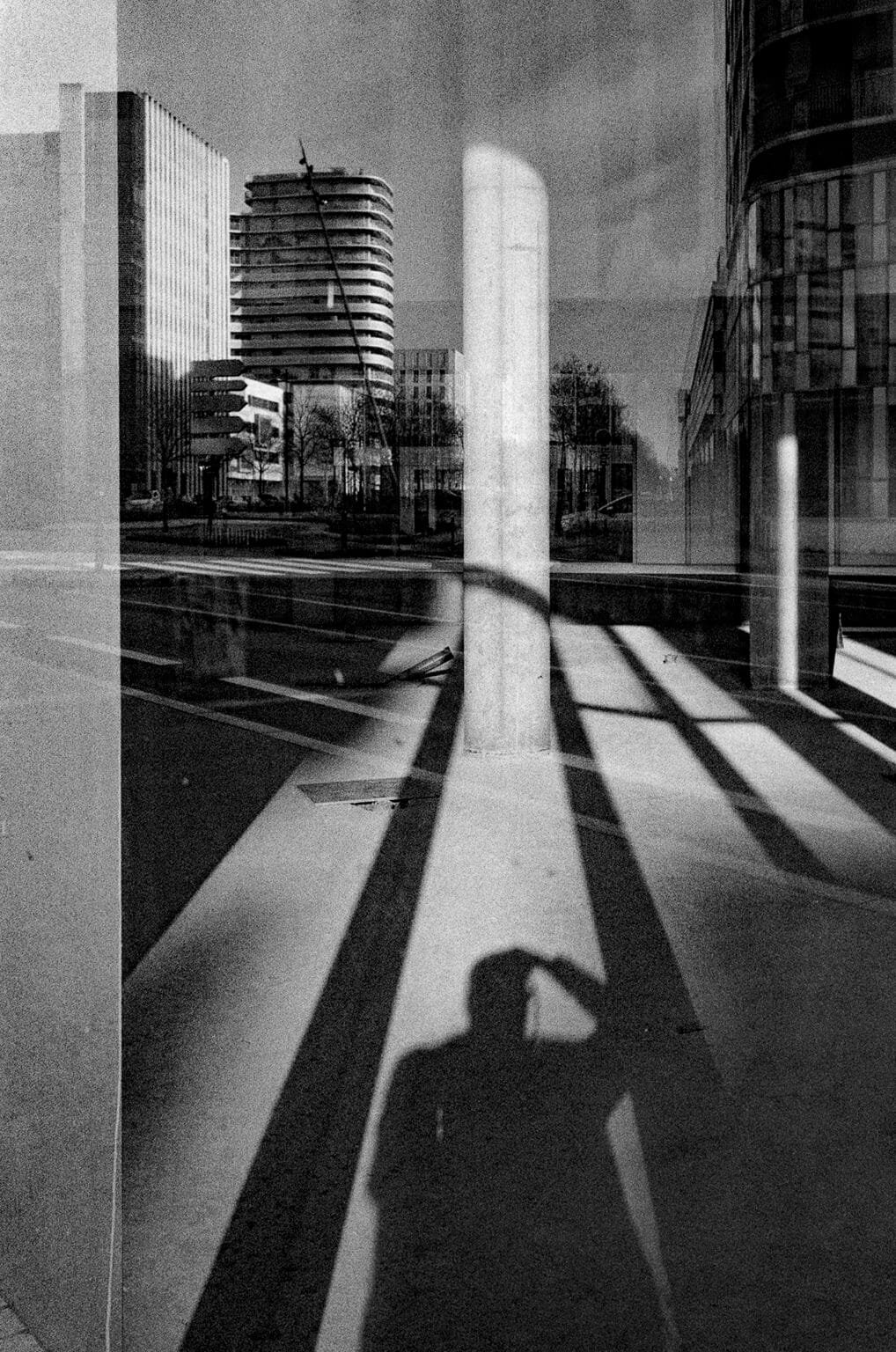 Reflet urbain dans une vitrine à Nantes, mêlant immeubles contemporains, poteau central et ombre du photographe au sol.