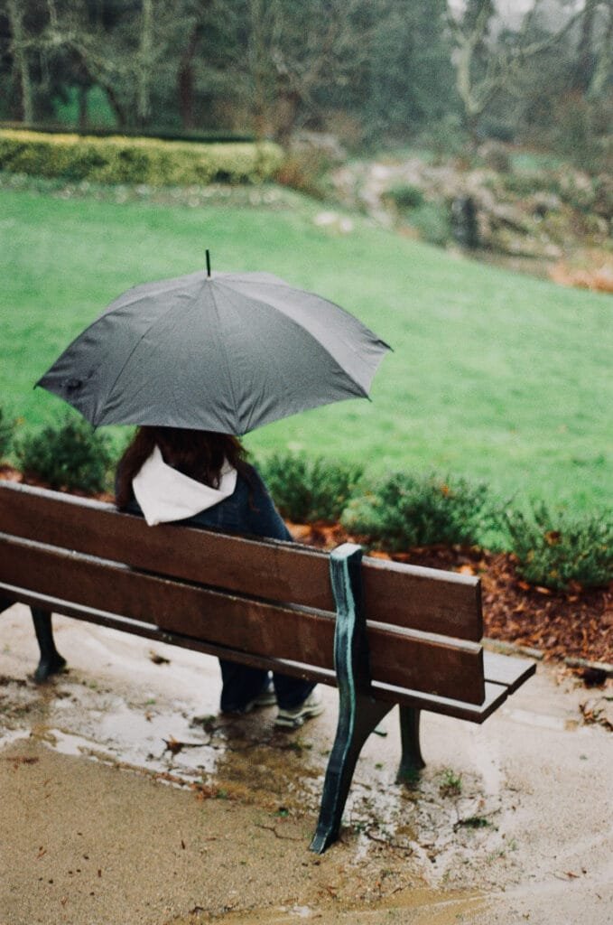 Personne assise sur un banc sous un parapluie dans un parc à Nantes par temps humide.