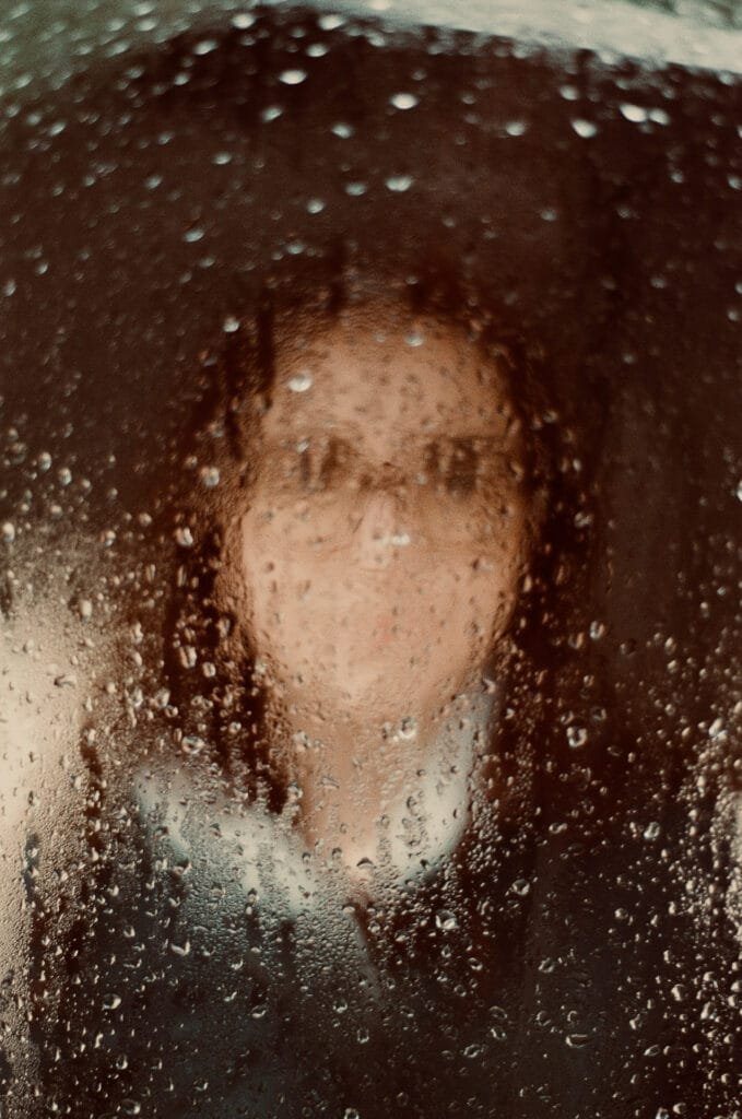 Silhouette d’un visage flou derrière une vitre couverte de gouttes de pluie.