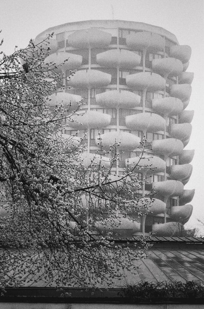 Photographie réalisée aux Choux de Créteil en mars 2026. Les tours d’habitation émergent dans une lumière diffuse et légèrement brumeuse, tandis que les arbres du premier plan dessinent des silhouettes sombres. Cette atmosphère adoucit les volumes des balcons et donne au bâtiment une présence presque irréelle. Nikon F3 HP chargé avec une pellicule noir et blanc Kentmere 400.