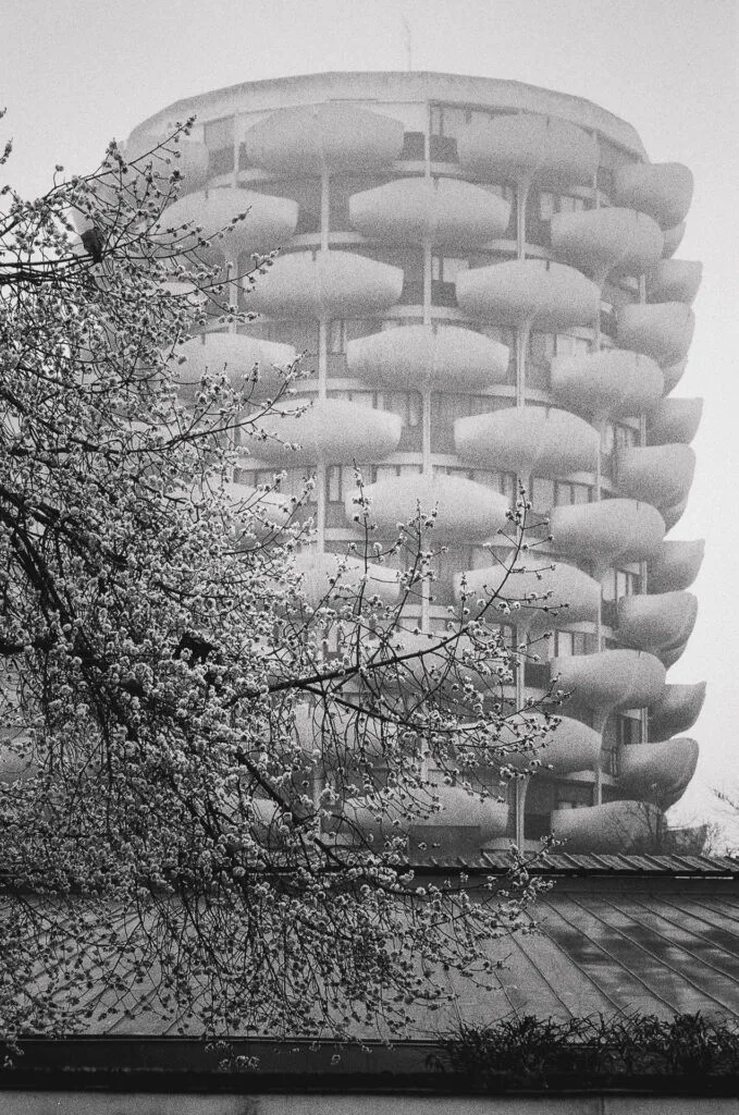 Photographie réalisée aux Choux de Créteil en mars 2026. Les tours d’habitation émergent dans une lumière diffuse et légèrement brumeuse, tandis que les arbres du premier plan dessinent des silhouettes sombres. Cette atmosphère adoucit les volumes des balcons et donne au bâtiment une présence presque irréelle. Nikon F3 HP chargé avec une pellicule noir et blanc Kentmere 400.