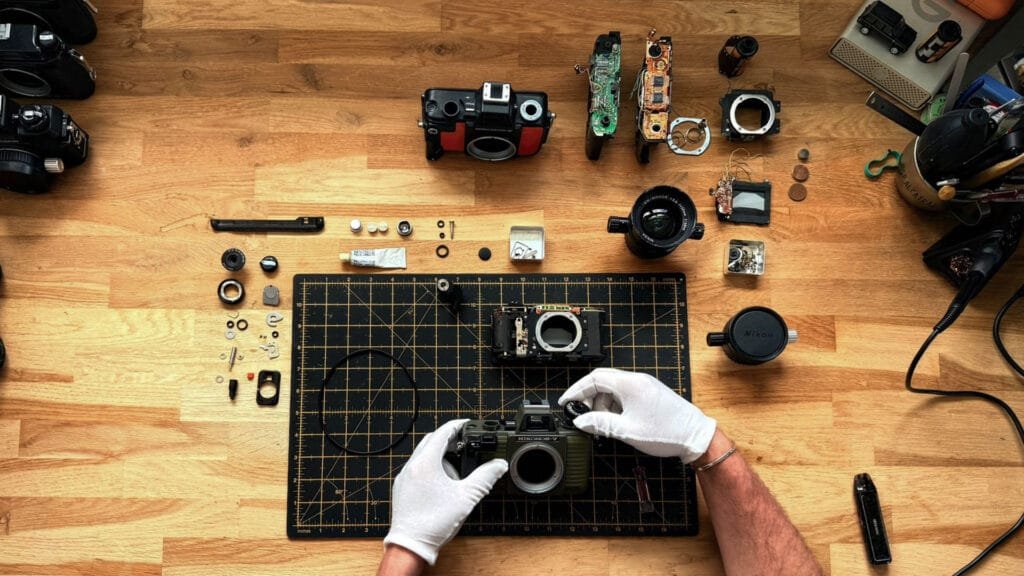 Démontage complet d'un Nikonos V vert olive, toutes les pièces disposées autour du boîtier sur un tapis de découpe, mains gantées d'un technicien en cours de révision.