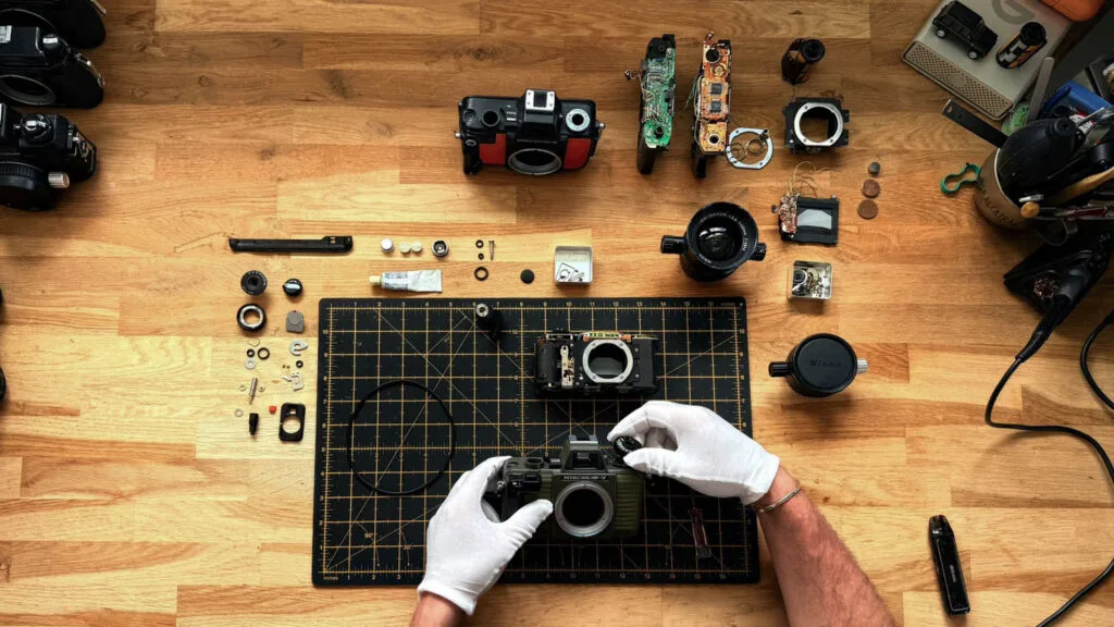 Démontage complet d'un Nikonos V vert olive, toutes les pièces disposées autour du boîtier sur un tapis de découpe, mains gantées d'un technicien en cours de révision.