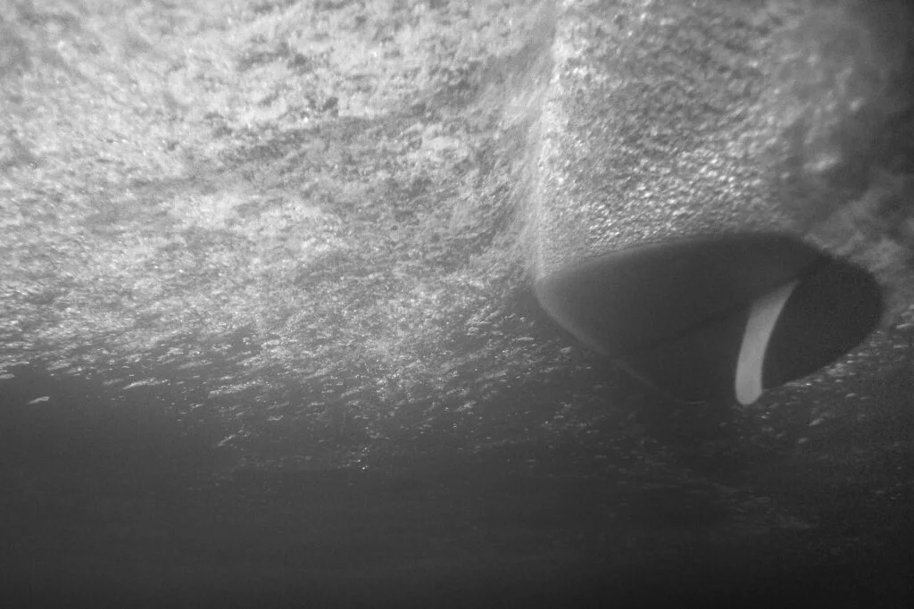Dérive de planche de surf vue depuis sous l'eau avec écume et bulles, photo argentique noir et blanc au Nikonos V - © YannB / yannb.net