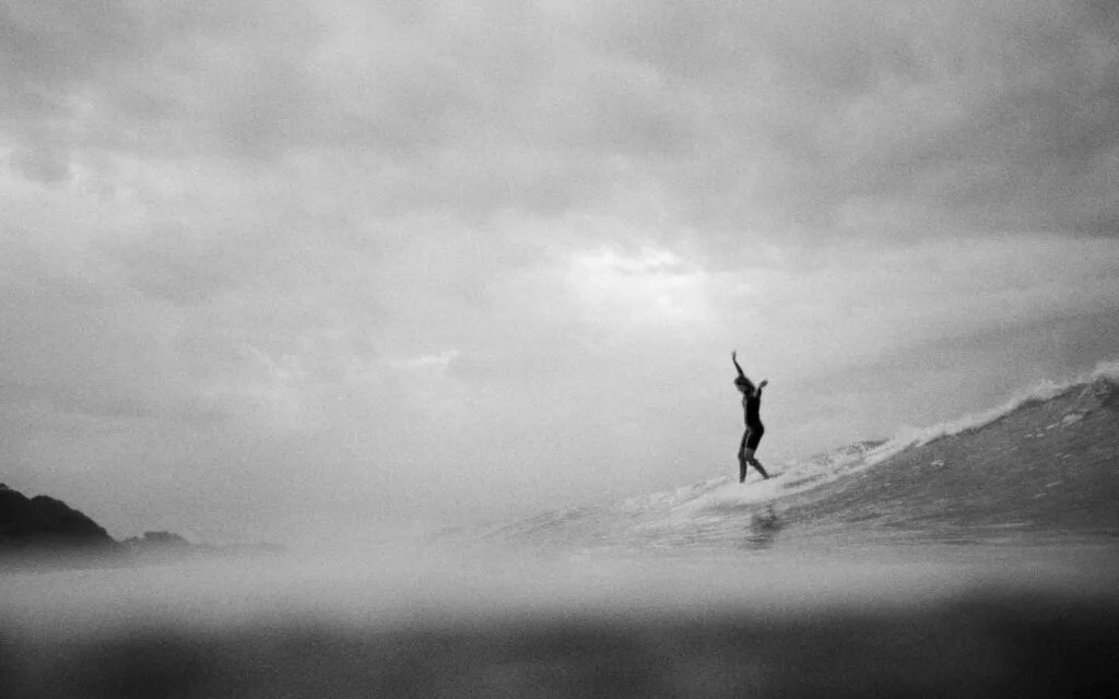 Surfeuse bras levés sur une vague sous un ciel nuageux et brumeux, photo argentique noir et blanc au Nikonos V - © YannB / yannb.net