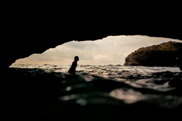 Surfeuse assise sur sa planche en silhouette dans l'encadrement d'une grotte marine, photo argentique couleur - © YannB / yannb.net