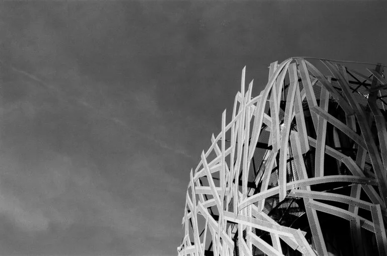 Contre-plongée sur une structure métallique tressée façon nid, ciel dégagé, argentique noir et blanc