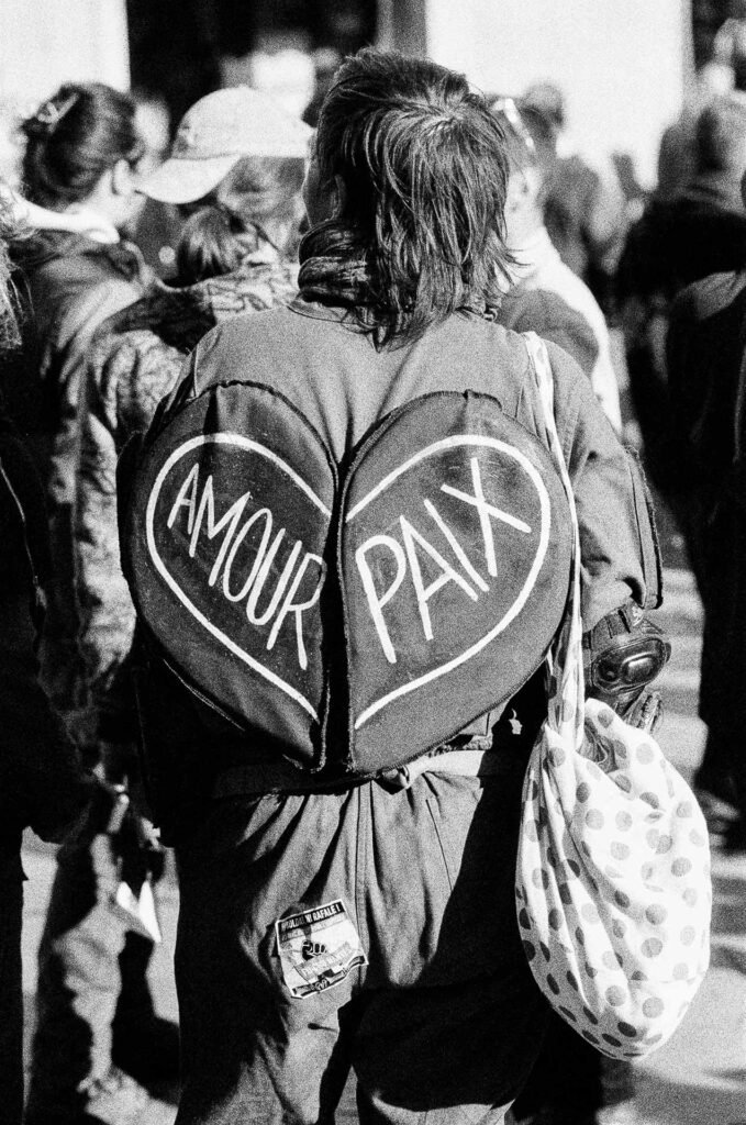 Manifestante vue de dos portant un grand cœur en tissu marqué « Amour Paix », sac à pois à l'épaule