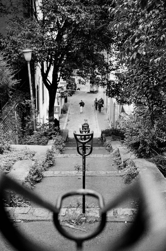 Escalier envahi par la végétation à Nantes, avec une rampe métallique floue au premier plan et des passants au loin