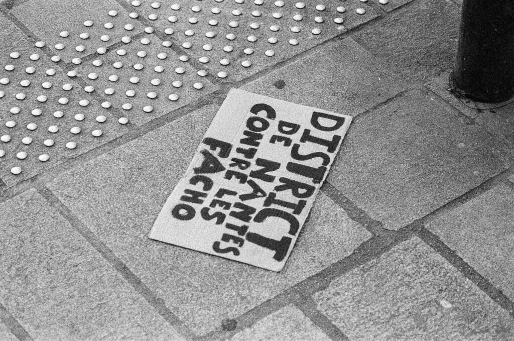 Pancarte en carton « District de Nantes contre les facho » abandonnée sur un trottoir à côté d'une bande podotactile