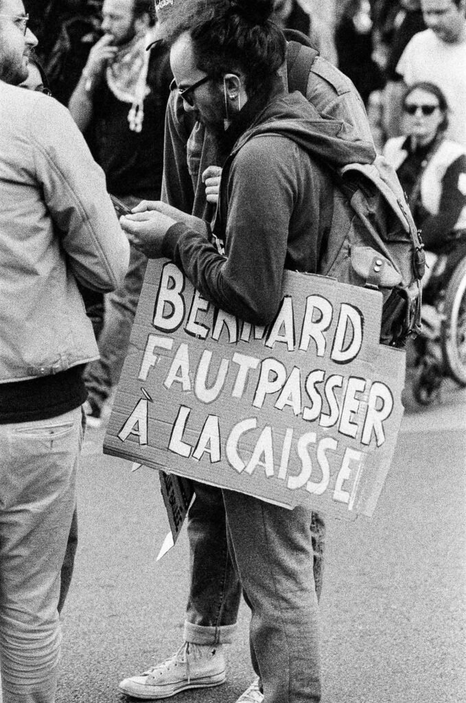 Manifestant consultant son téléphone, une pancarte « Bernard faut passer à la caisse » portée contre son sac à dos