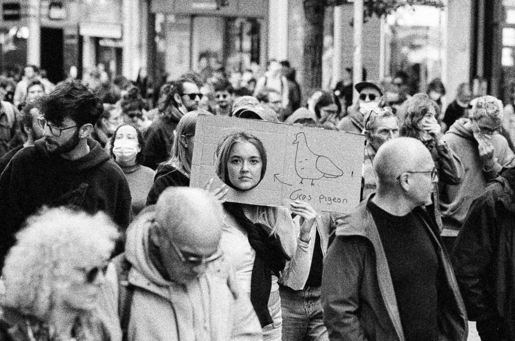 Manifestante passant son visage dans le trou d'une pancarte en carton où est dessiné un pigeon, au milieu de la foule à Nantes