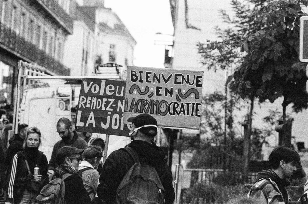 Pancartes « Bienvenue en lacrymocratie » et « Voleurs rendez-nous la joie » au-dessus d'un groupe de manifestants dans une rue arborée de Nantes