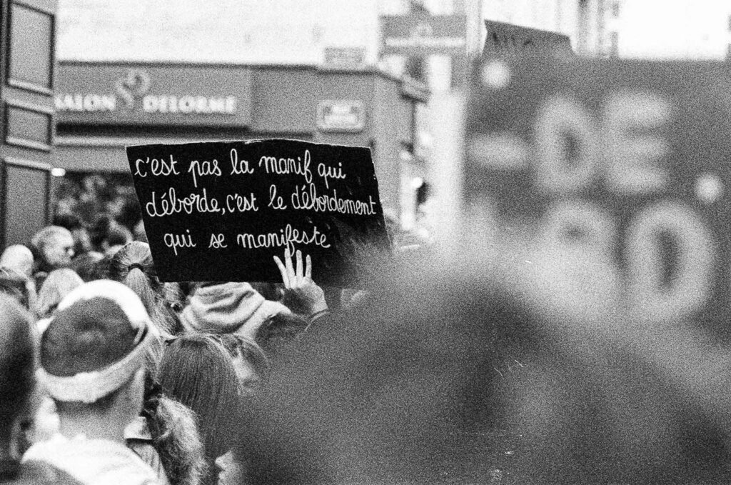 Pancarte noire écrite à la craie « C'est pas la manif qui déborde, c'est le débordement qui se manifeste » au-dessus de la foule, premier plan flou