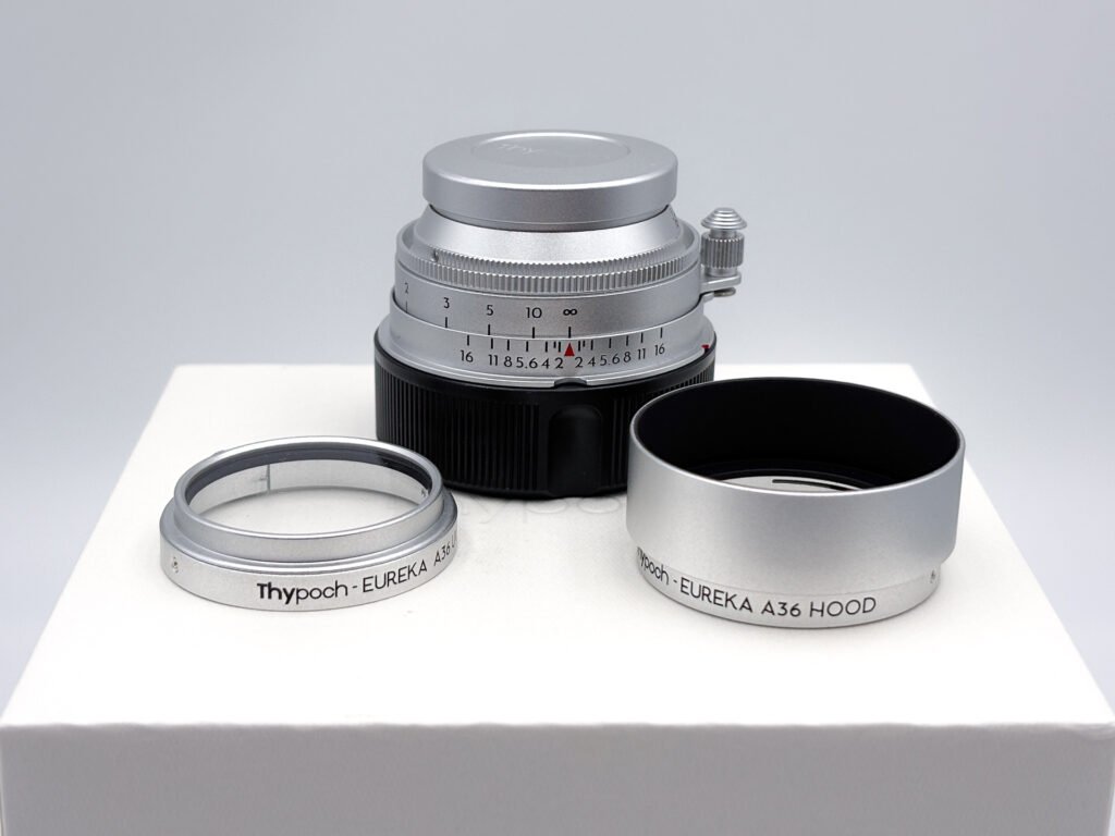 Objectif Thypoch Eureka 50mm f/2 en aluminium rentré avec ses bouchons, accompagné de son filtre UV et de son pare-soleil gravés « Thypoch - EUREKA A36 », posés sur la boîte d’origine