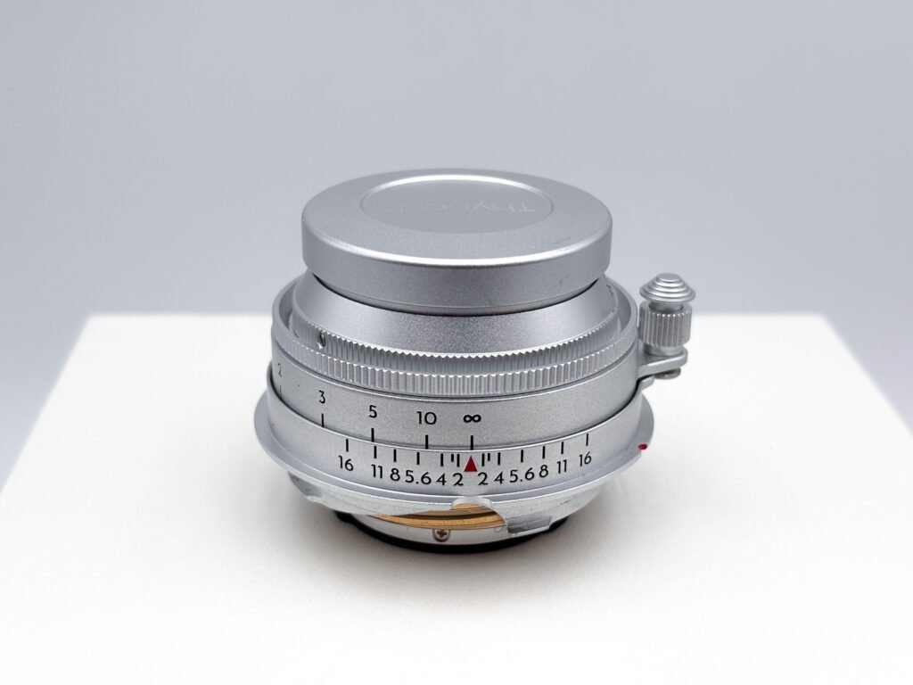 Objectif Thypoch Eureka 50mm f/2 en aluminium posé seul sur un socle blanc, rentré avec son bouchon avant, montrant son échelle des distances, son échelle de profondeur de champ et la came de couplage du télémètre au niveau de la monture M