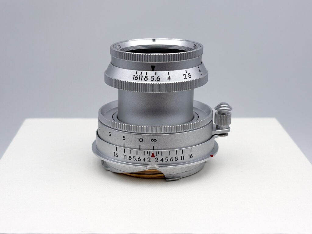 Objectif Thypoch Eureka 50mm f/2 en aluminium posé seul sur un socle blanc, en position sortie, laissant voir le tube coulissant entre la bague de mise au point et la bague d’ouverture réglée sur f/5.6