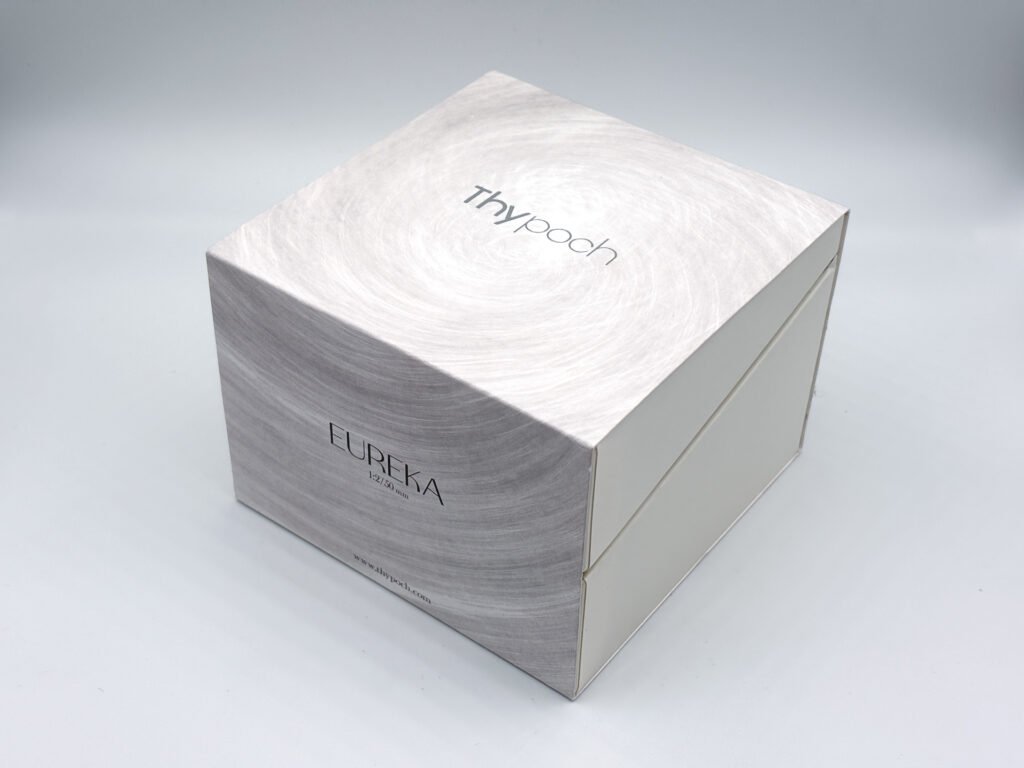 Boîte fermée de l’objectif Thypoch Eureka 50mm f/2, en carton gris clair à motif brossé circulaire, avec le logo Thypoch argenté sur le dessus et l’inscription EUREKA 1:2/50 mm sur la face avant