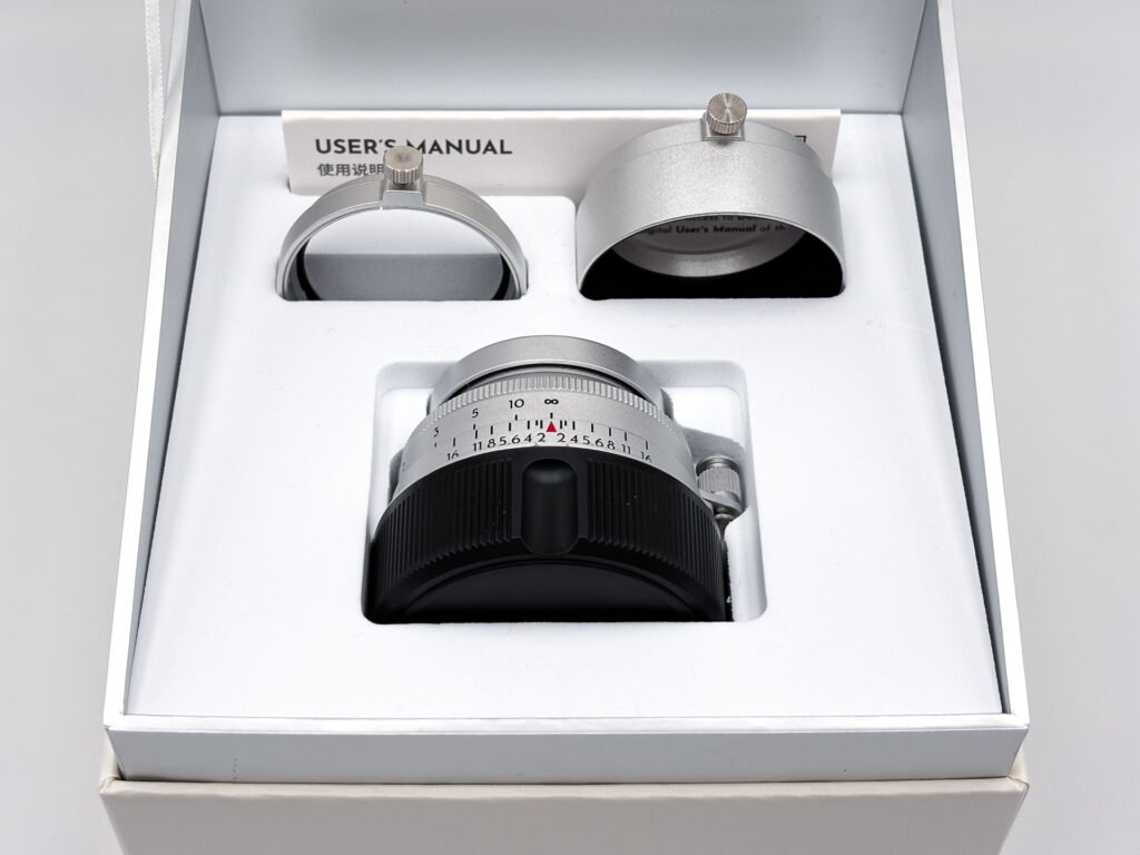 Objectif Thypoch Eureka 50mm f/2 en aluminium rangé dans sa boîte avec son échelle des distances visible, entouré de son filtre UV et de son pare-soleil à vis de serrage