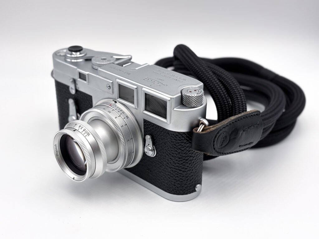 Leica M3 chromé de 1958 vu de trois quarts, équipé de l’objectif rétractable Thypoch Eureka 50mm f/2 en version aluminium, en position sortie, avec sa courroie noire