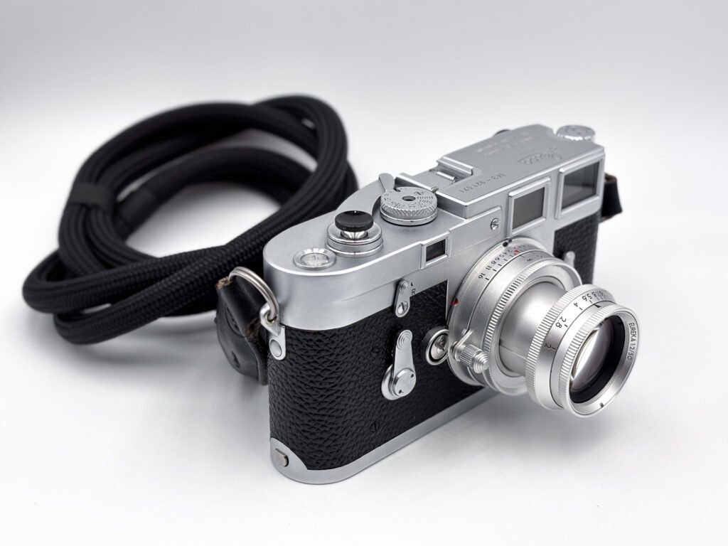 Leica M3 chromé de 1958 vu de trois quarts en légère plongée, équipé de l’objectif Thypoch Eureka 50mm f/2 en aluminium en position sortie, avec sa courroie noire enroulée à l’arrière